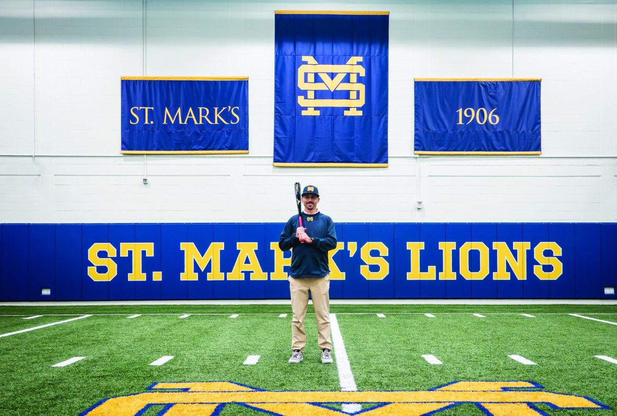 Head baseball Coach Dennis Kelly  stands next to the batting cages in the fieldhouse, bat in hand. Along with coaching baseball, Kelly also coaches P.E. as well as various other sports on campus.