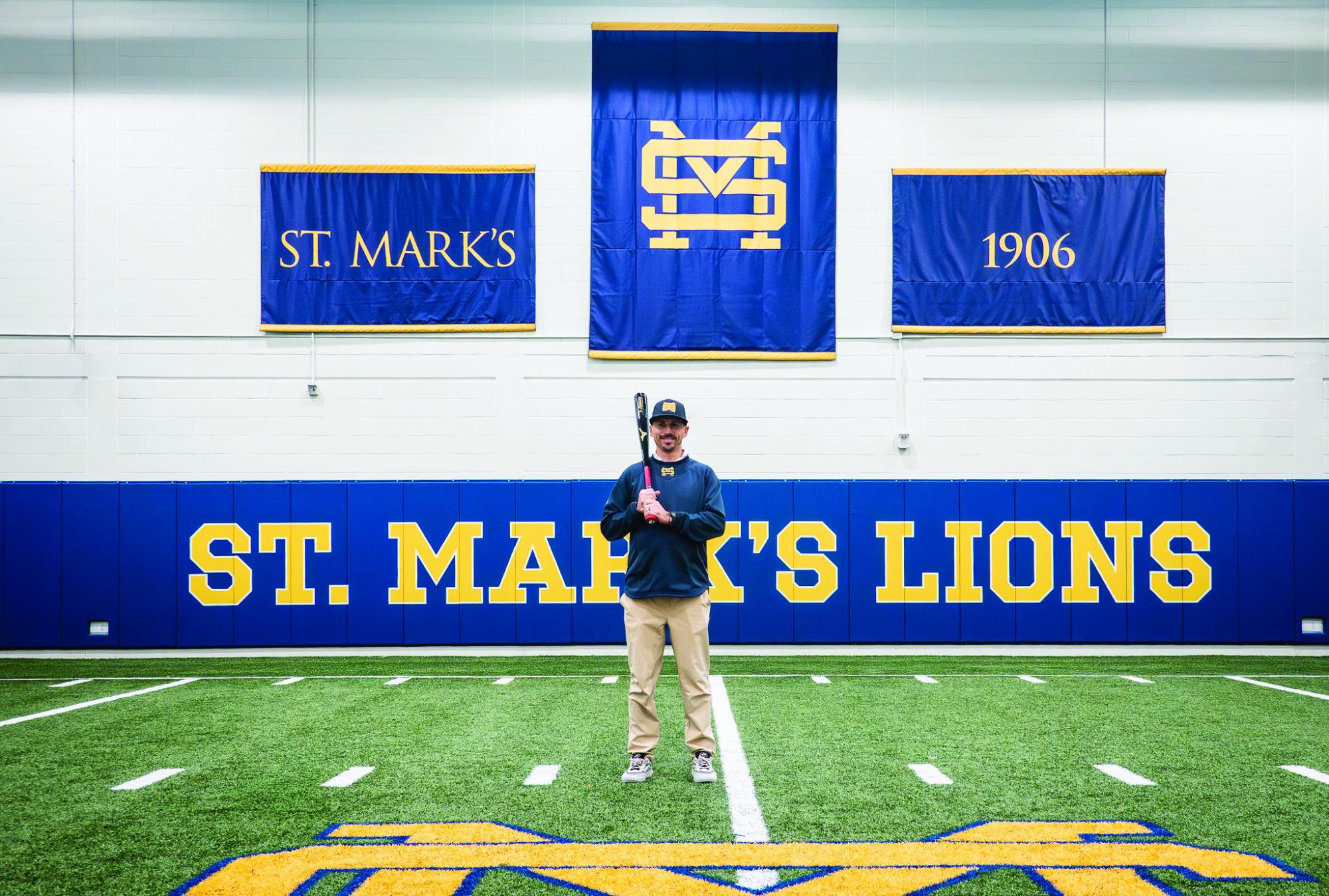 Head baseball Coach Dennis Kelly  stands next to the batting cages in the fieldhouse, bat in hand. Along with coaching baseball, Kelly also coaches P.E. as well as various other sports on campus.
